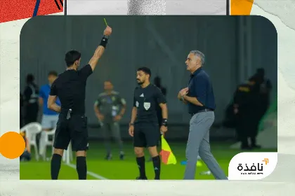 Soccer referee raises a yellow card toward a coach on the sideline as players watch on a green field.