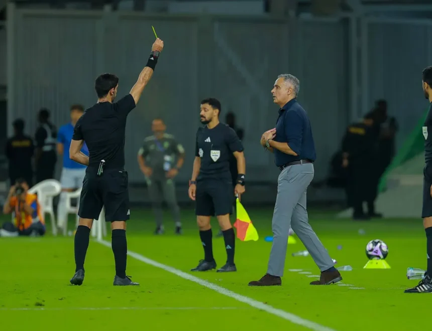 Soccer referee raises a yellow card toward a coach on the sideline as players watch on a green field.